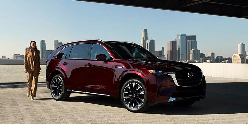 woman walking up to a burgundy Mazda CX-90 with a city skyline in the distance