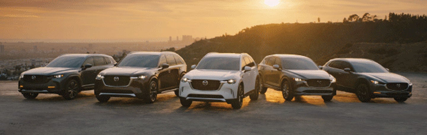 Five Mazda SUVs (CX-50, CX-90, CX-90 PHEV, CX-5, CX-30) parked side-by-side at sunset over a city.
